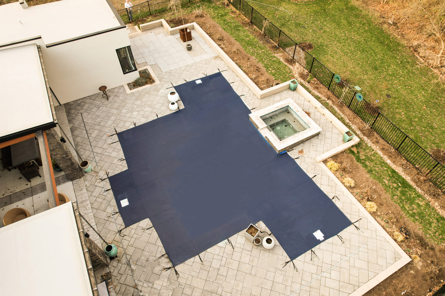 Overhead view of a dark blue mesh safety cover secured with straps over a custom-shaped in-ground swimming pool, adjacent to a raised square spa, surrounded by a paved patio, modern home, and fenced lawn.