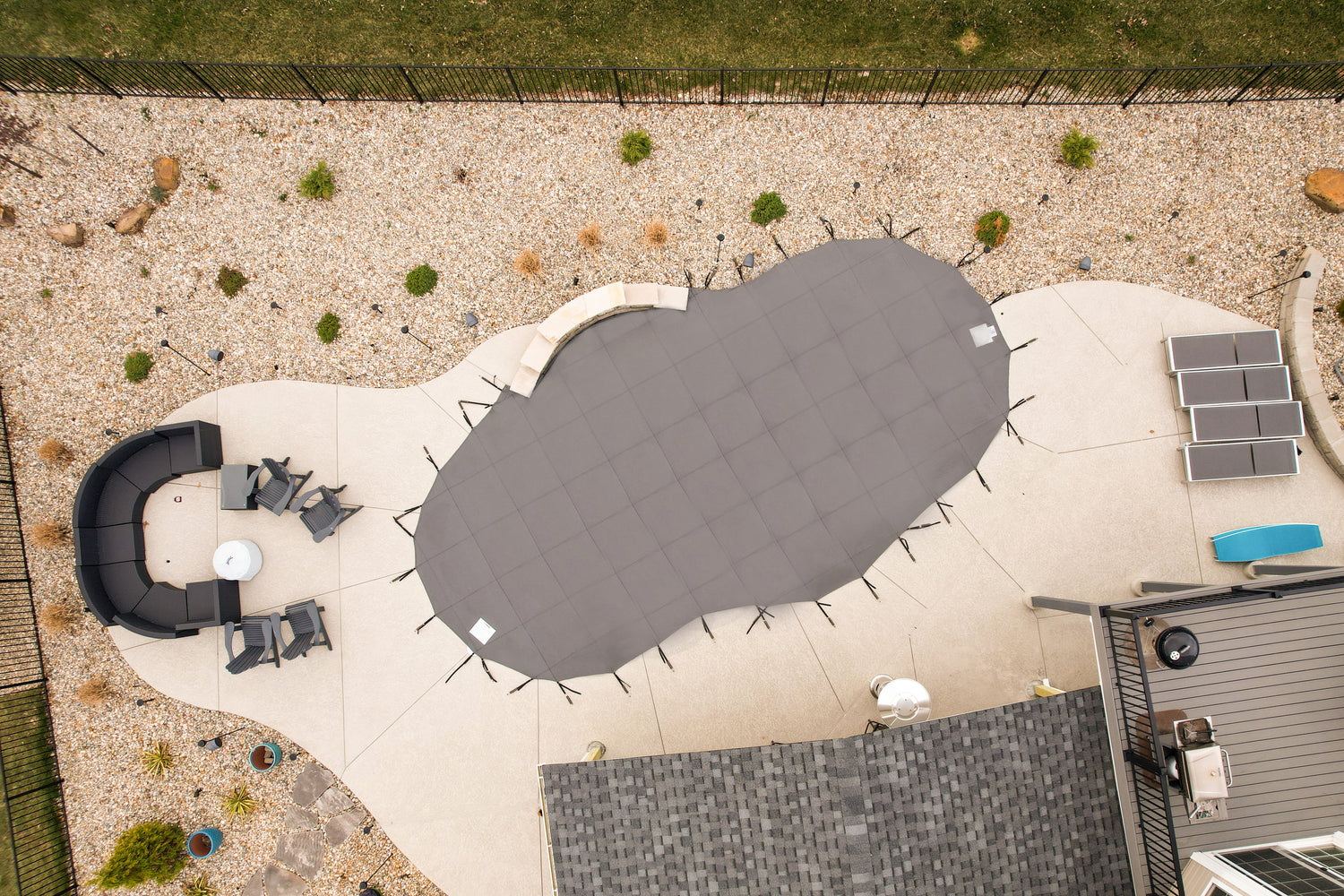 Overhead view of a gray mesh safety cover secured with straps over a freeform in-ground swimming pool, surrounded by a concrete patio with outdoor seating, landscaping stones, and a fenced yard.