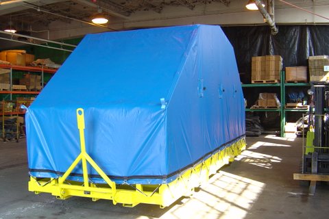 Large industrial cargo covered with a fitted blue tarp, secured on a yellow metal platform inside a warehouse. The tarp forms a tent-like shape with tie-down points along the sides, and shelving, pallets, and forklifts are visible in the background under overhead lighting.