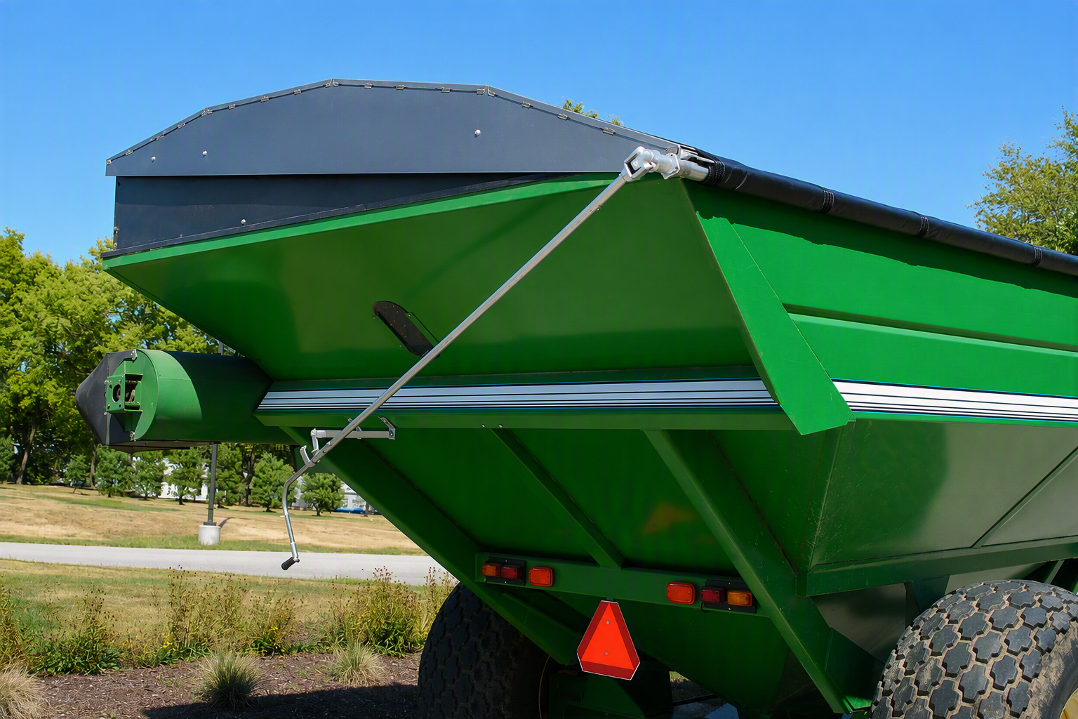 A green agricultural spreader or hopper trailer shown outdoors on a sunny day, with trees and blue sky in the background. The rear body of the trailer is visible, including a dark gray top cover, metal support ropes, wheels, and a red triangular reflector.
