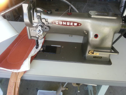 Industrial CONSEW brand sewing machine mounted on a worktable, shown stitching thick brown and white material. The heavy-duty machine has metal components, visible thread guides, and a needle positioned over layered fabric, indicating contract or industrial sewing work in a workshop setting.