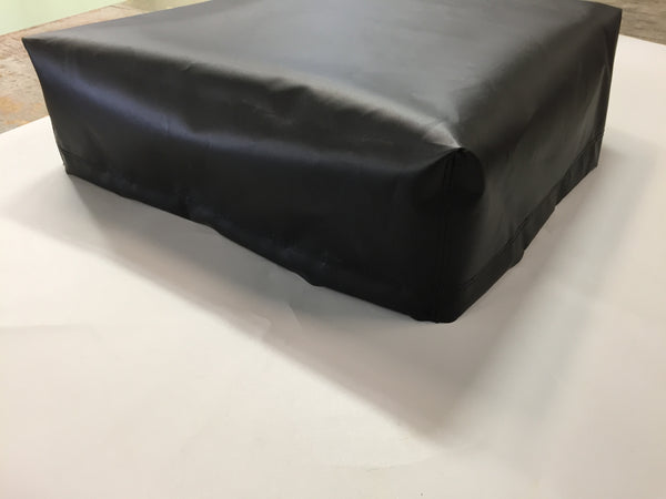 Black rectangular vinyl box cover fitted snugly over a low-profile object, with smooth surfaces and slightly rounded corners, resting on a white tabletop in an indoor workspace.
