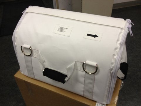 White rectangular protective cover made of heavy-duty fabric with a flap top, metal D-rings, and a small black handle on the front. The cover rests on a cardboard box indoors, with a small label and an arrow symbol visible on the front panel.
