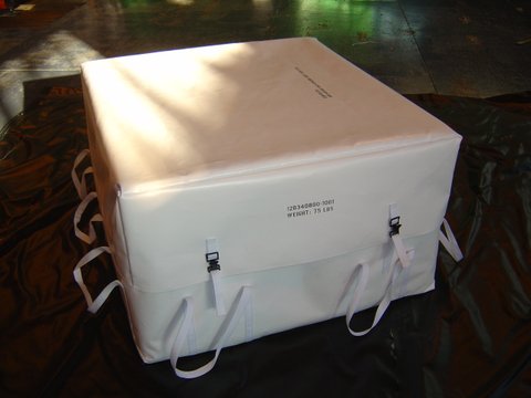 White rectangular soft-sided equipment cover with a fitted lid, webbing handles along the sides, and two front buckles. Printed identification text is visible on the front panel. The cover rests on a dark tarp surface in an indoor industrial setting with light reflecting across the top.