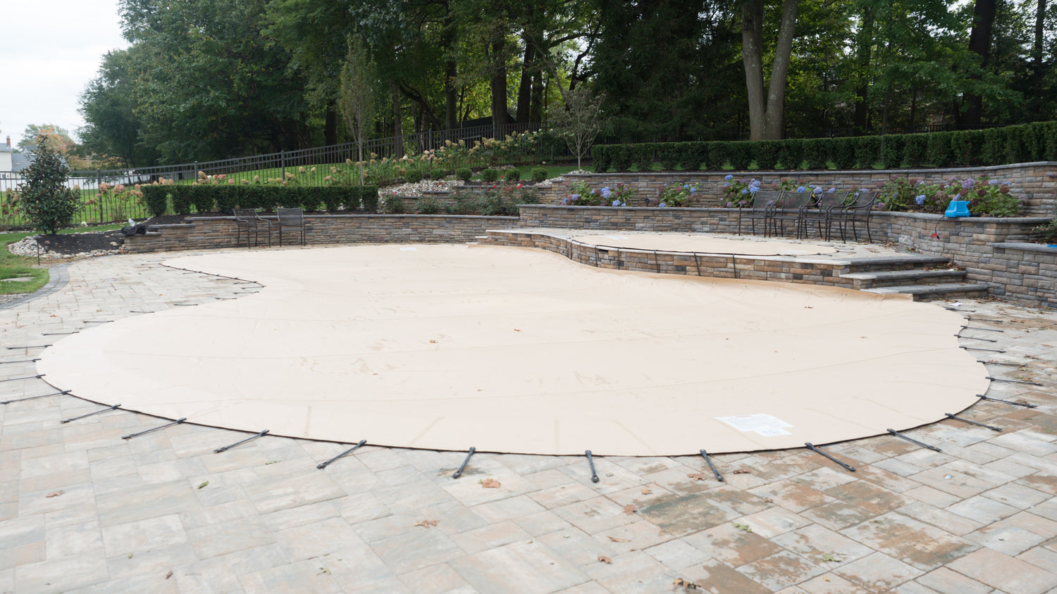 Tan safety cover secured with straps over a freeform in-ground swimming pool, surrounded by a paved patio, stone retaining walls, landscaped garden beds, and trees in the background.