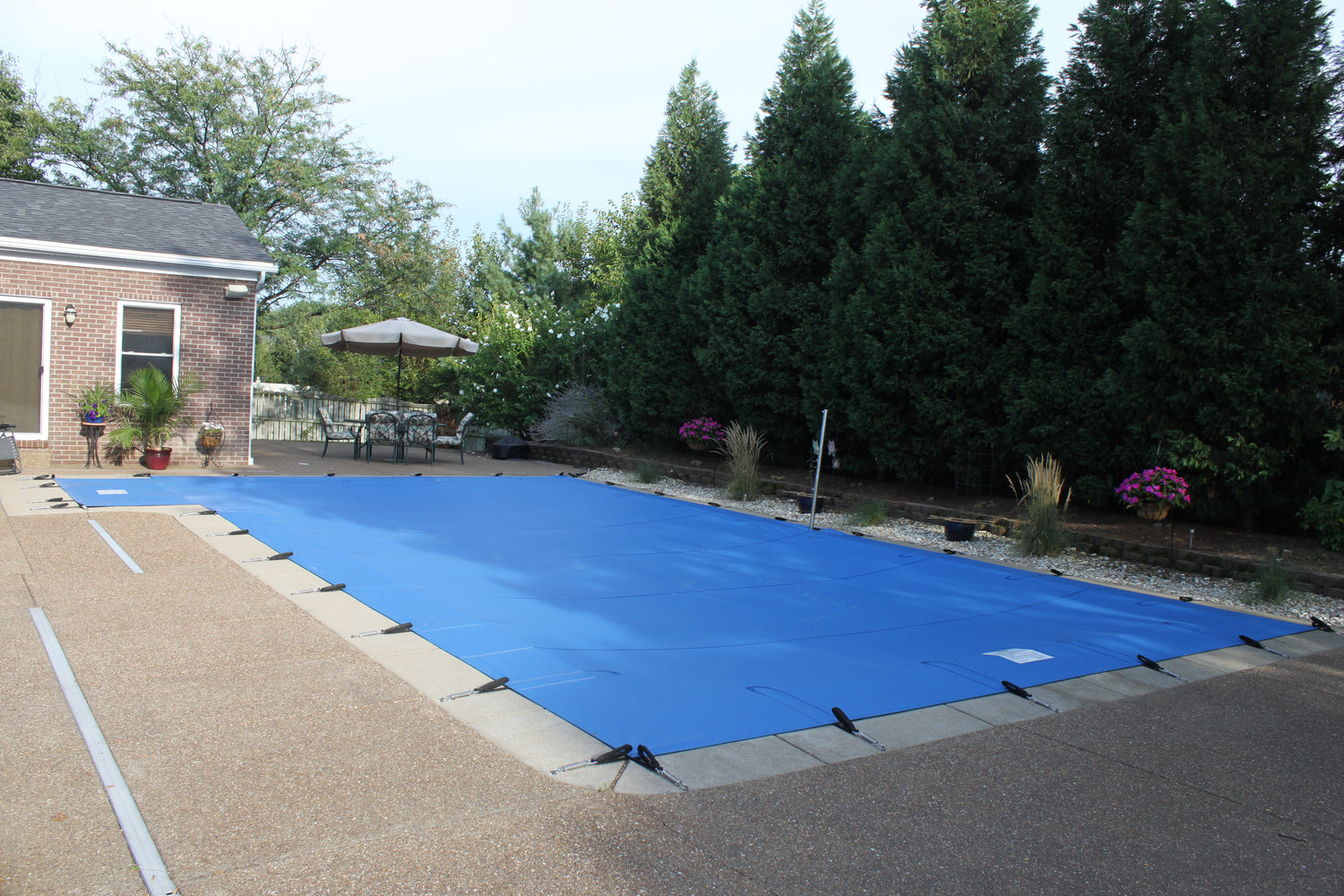 Blue safety cover secured with straps over an in-ground rectangular swimming pool, set within a concrete patio beside a brick house, with outdoor seating, an umbrella, and tall evergreen trees lining the yard.