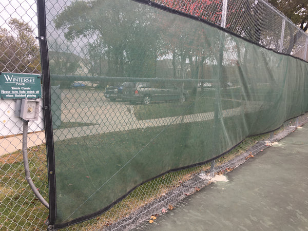green mesh windscreen on tall chain link fence dividing a court and a grass area with sidewalk. Fence includes sign that says "Winterset park tennis courts please turn light switch off when finished playing"